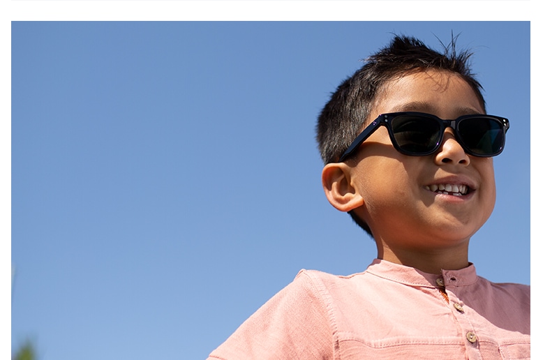 Young boy with short brown hair standing outside wearing Wayfarer-style black acetate sunglasses #4433121 with photochromic lenses and a pink short-sleeve pocket tee.