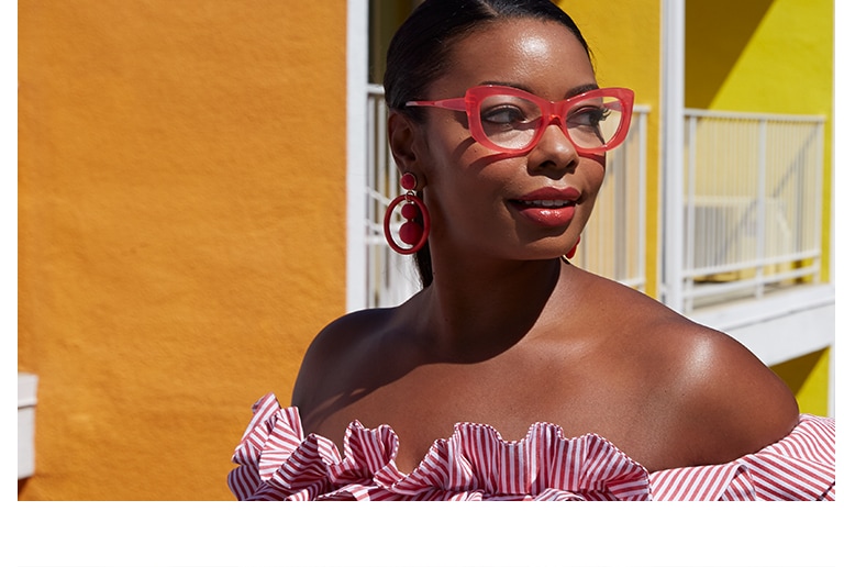 African-American woman with hair pulled back wearing bold candy pink cat-eye glasses #4438119 and off-the-shoulder pink-and-white striped ruffle top standing in front of vibrant yellow-and-orange façade of Saguaro Hotel in Palm Springs.