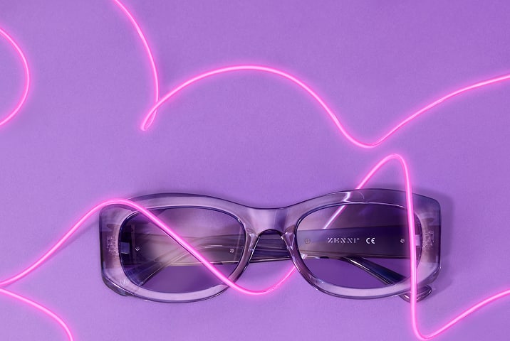 A pair of translucent purple retro-style sunglasses lies on a purple surface, surrounded by looping neon pink light streaks.