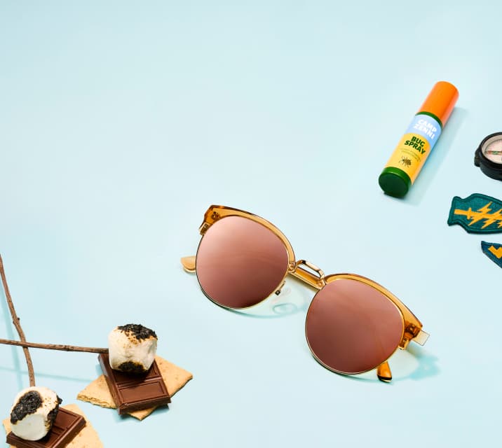 Round browline sunglasses with translucent amber frames and rose gold reflective lenses resting on a light blue background, surrounded by camping-themed items like a s'more, bug spray, a compass, and patches.