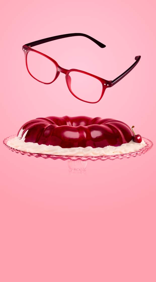 Red frame glasses floating above a cherry-red gelatin ring mold with cream on a glass cake stand, set against a pink background.