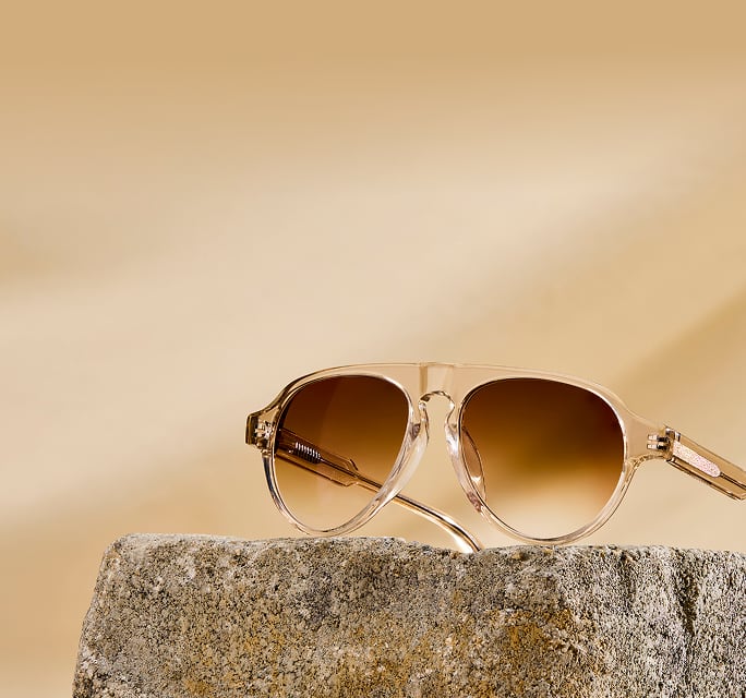 Light brown aviator sunglasses with brown gradient lenses, on a beige stone background.