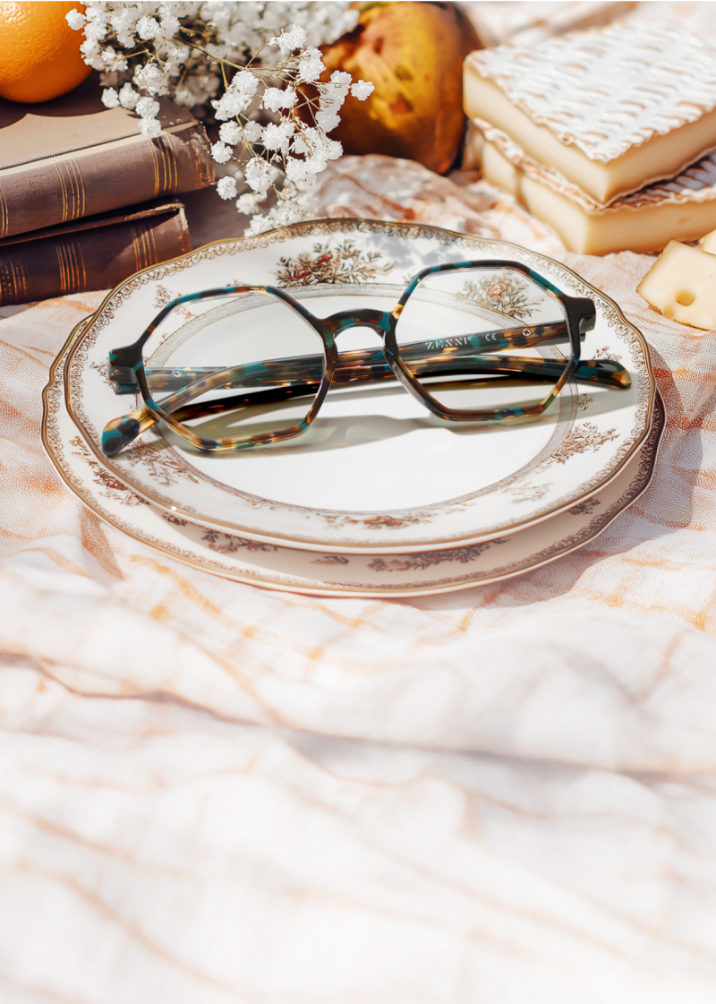 Geometric brown and blue tortoiseshell glasses resting on an antique china plate, on a pale pink picnic blanket with vintage books, brie cheese, and white flowers nearby.