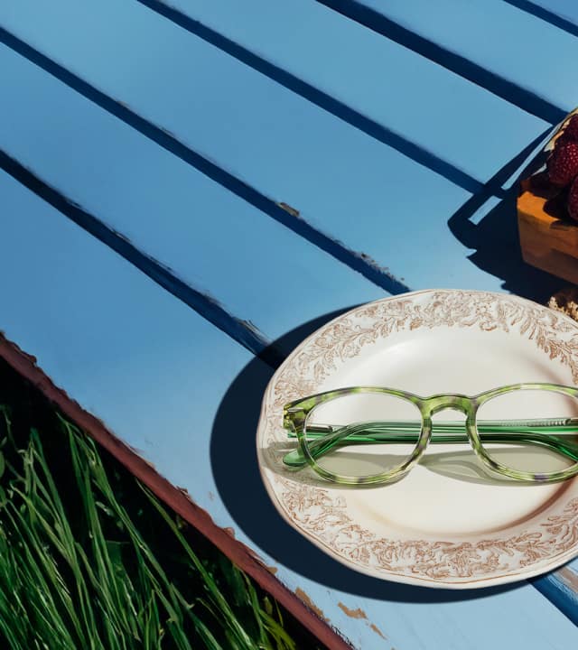 Round green tortoiseshell glasses resting on an antique china plate, sitting on a blue painted wood picnic bench beside a small basket filled with raspberries and white daisies.