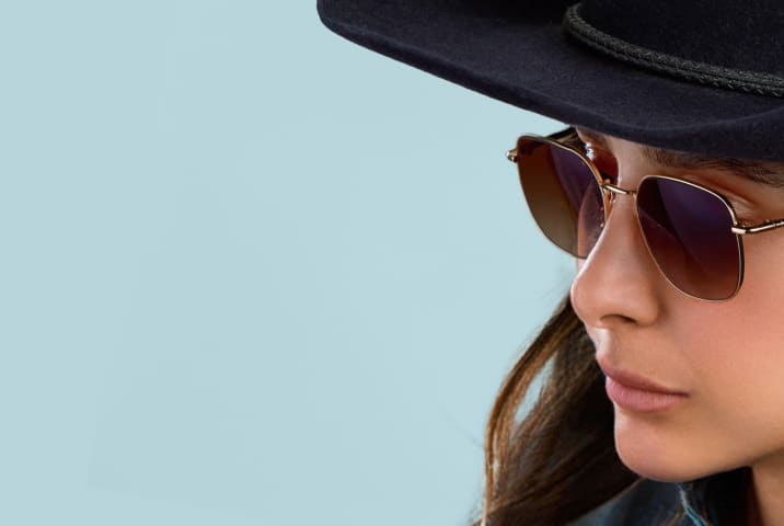 Close-up of a person wearing a black cowboy hat and gold wireframe sunglasses with dark brown gradient lenses, set against a light blue background.