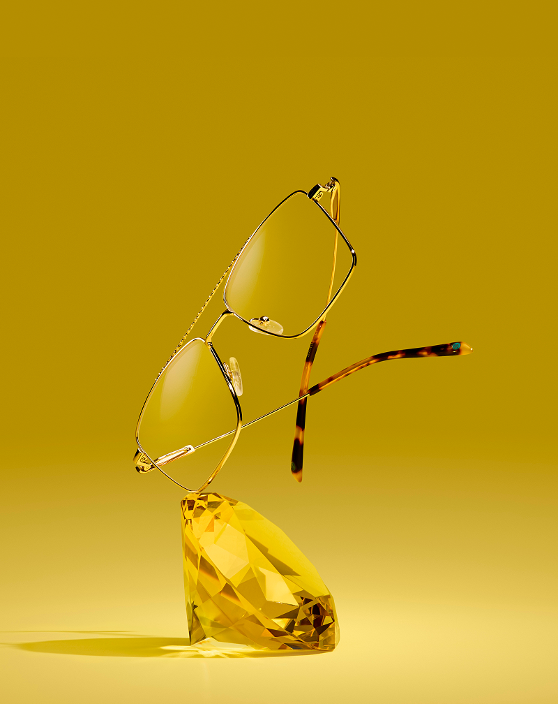 Gold aviator glasses resting on a giant topaz gem, on a yellow background.