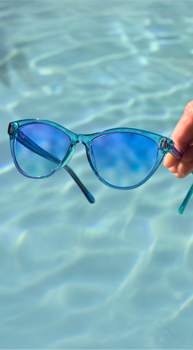 Hand holding Zenni translucent blue cat-eye sunglasses over shimmering pool water.
