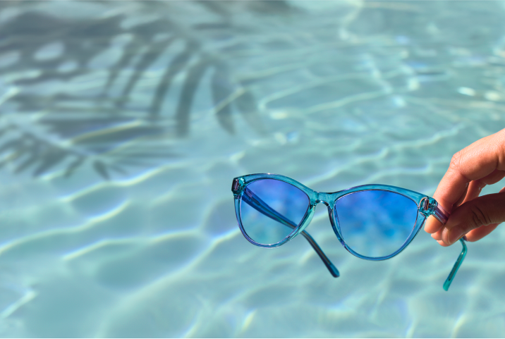 Hand holding Zenni translucent blue cat-eye sunglasses over shimmering pool water.