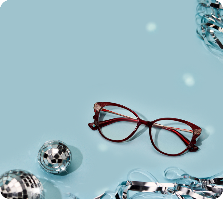 A pair of red, glitter-accented cat-eye glasses resting on a light blue surface next to two small disco balls and silver tinsel.