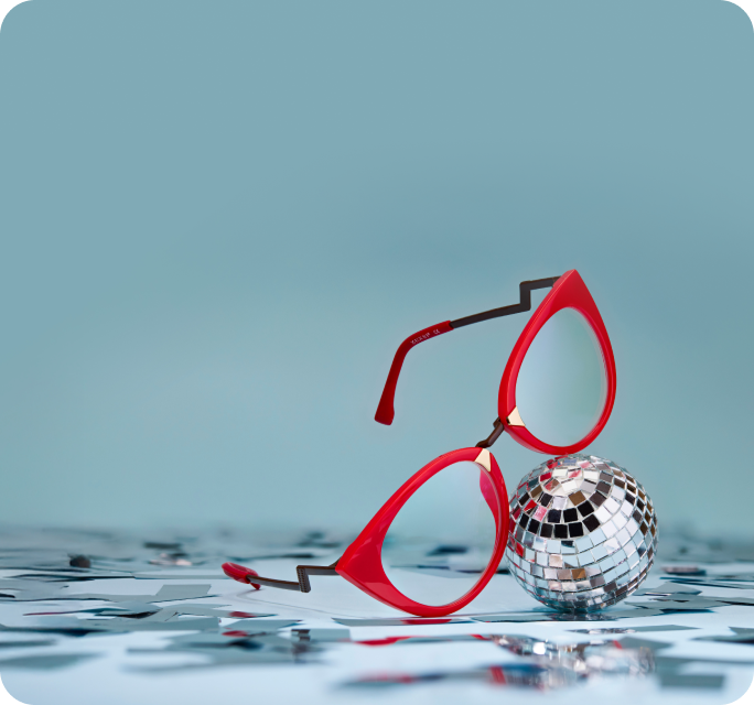 A close-up of a pair of angular red glasses with black zig-zag arms, leaning against a small disco ball on a light blue surface with silver confetti.