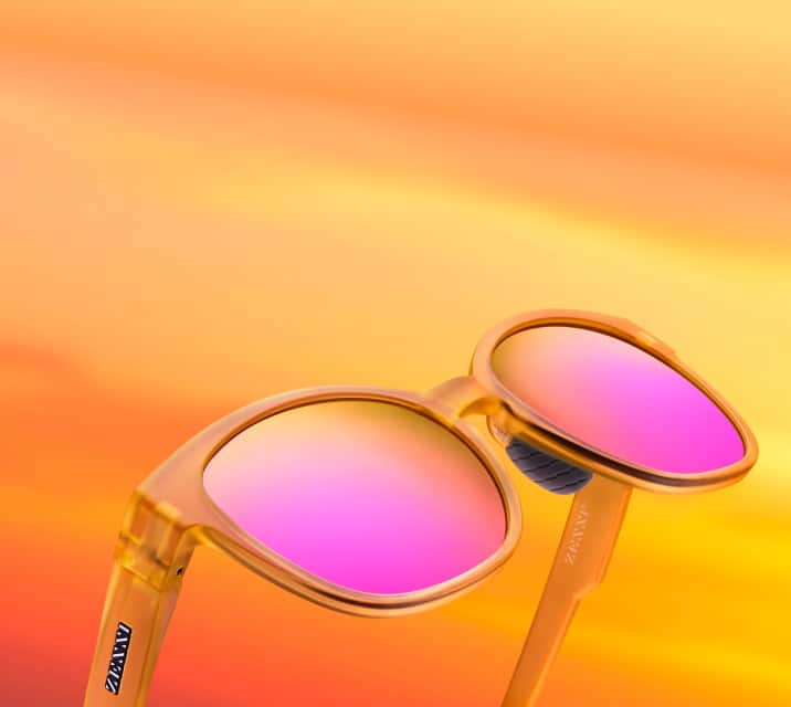 Translucent yellow-orange sunglasses with vibrant pink reflective mirror lenses, set against a warm orange and yellow gradient background.