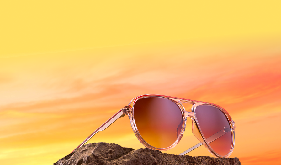 Clear pink aviator sunglasses with warm amber gradient lenses resting on a rough rock surface against a vibrant orange and yellow sunset sky.
