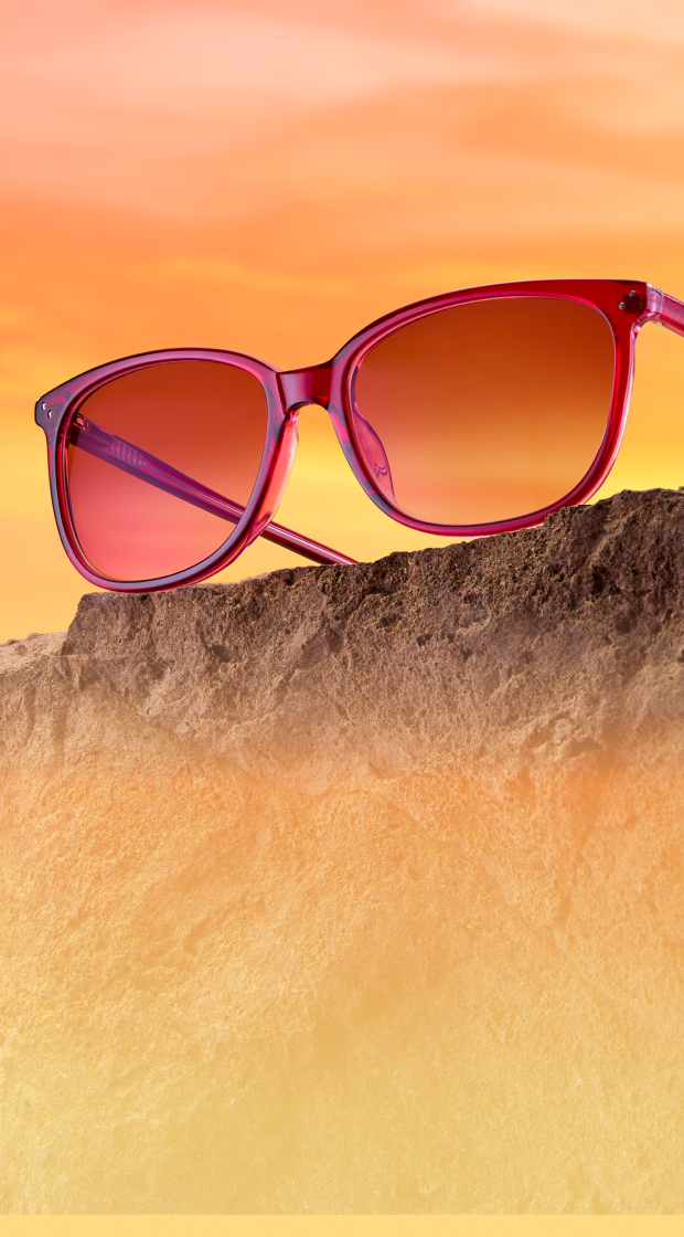 Translucent magenta-pink sunglasses with warm gradient lenses resting on a rough rock formation against a vibrant orange sunset sky.
