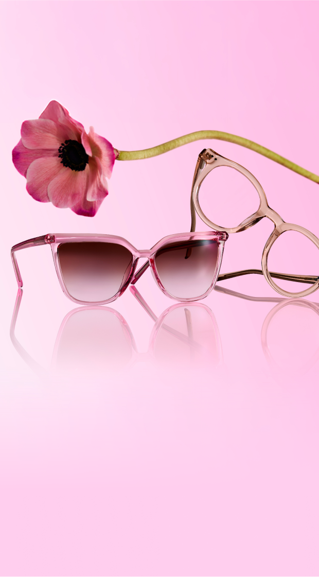 Two pairs of pink translucent eyeglasses—one cat-eye with gradient lenses and one round with clear lenses—are displayed on a reflective surface against a soft pink background, with a large pink flower arched gracefully above them.