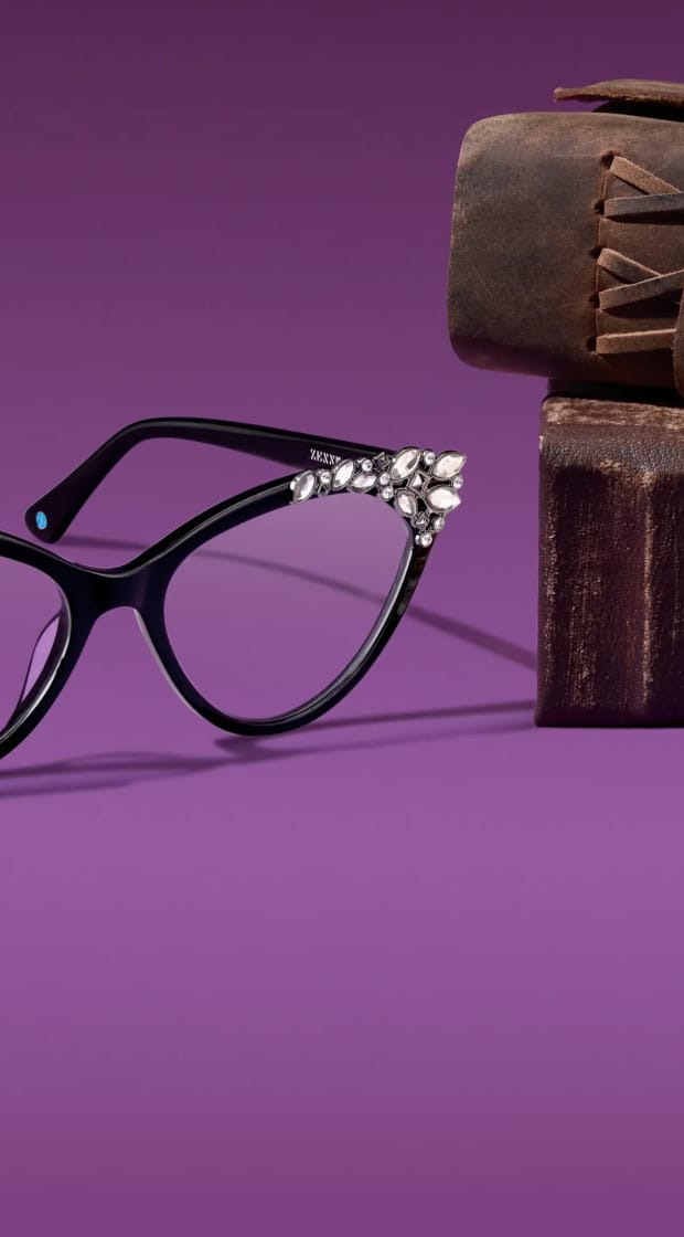 Black cat-eye eyeglasses adorned with sparkling crystals on the outer corners resting next to stacked vintage leather-bound books on a purple background.