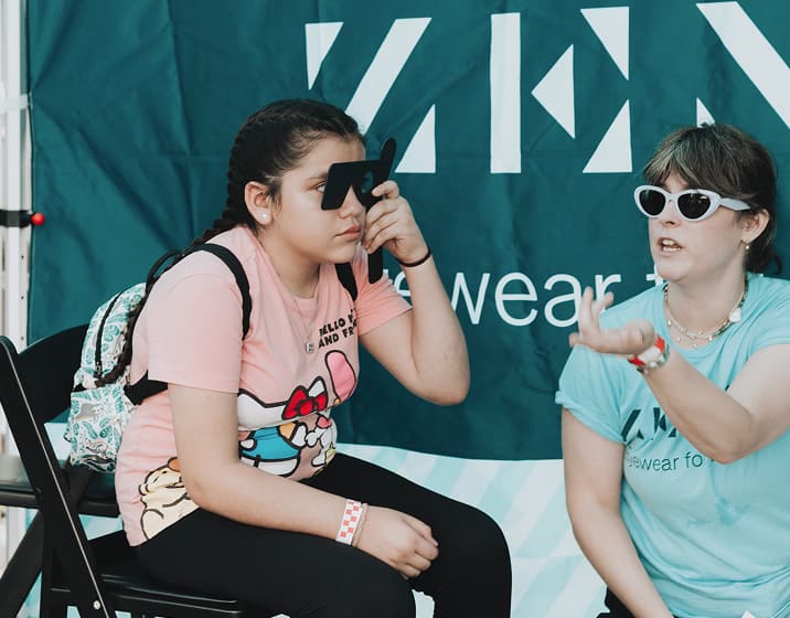 A young girl wearing a Hello Kitty t-shirt sitting on a chair and holding an eye occluder over her eye during a vision screening, while a woman in white sunglasses and a light blue t-shirt talks to her in front of a green Zenni banner.
