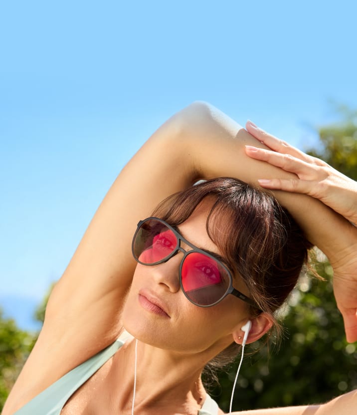 Close-up of a young woman with brown hair stretching her arms behind her head outdoors, wearing white wired earbuds and black aviator-style sunglasses with vibrant, reflective red lenses.