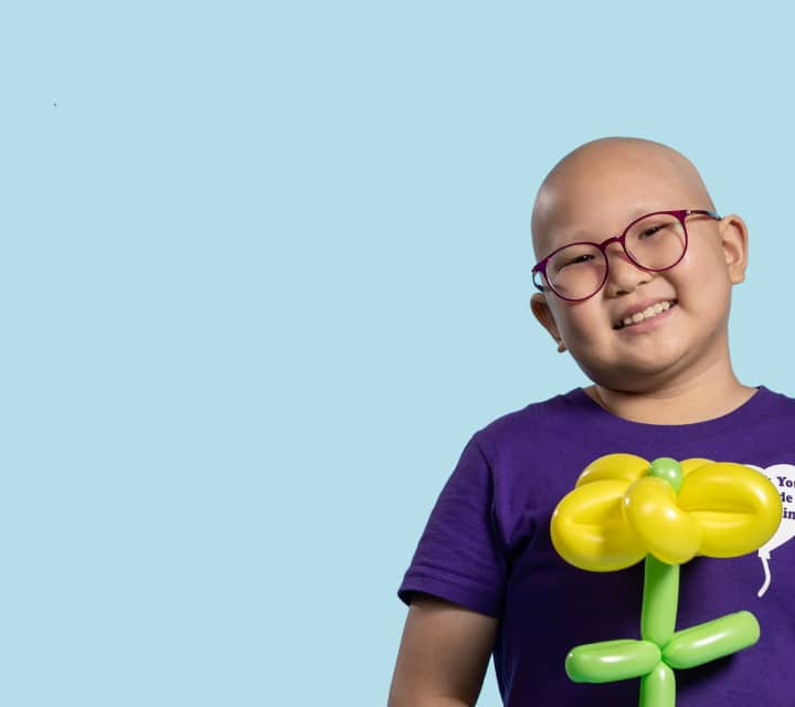 A smiling child wearing round, pink glasses and a purple shirt with a white heart graphic. They are holding a yellow and green balloon shaped like a flower. The background is a solid light blue, creating a cheerful and uplifting mood.