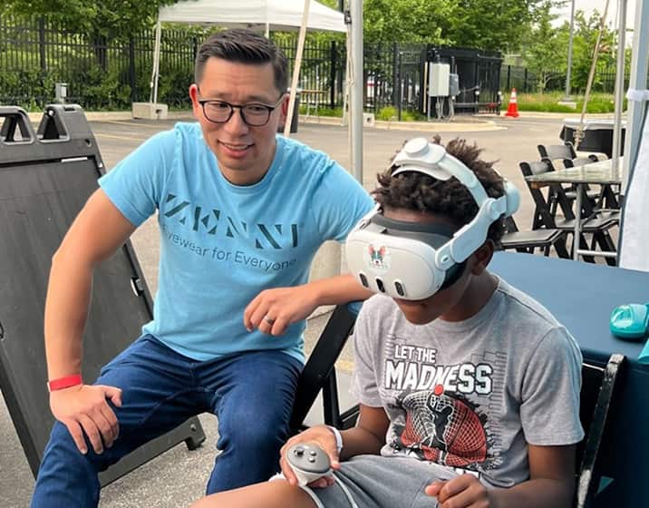 An adult man in glasses and a Zenni Volunteer t-shirt helps as a young boy, wearing a virtual-reality headset at a Zenni Vision Screening Event.