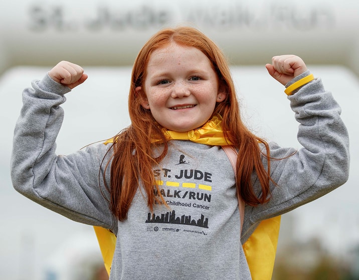 Smiling young girl in a St. Jude Walk/Run event, wearing a gray event sweatshirt and yellow cape, raising her fists in the air.