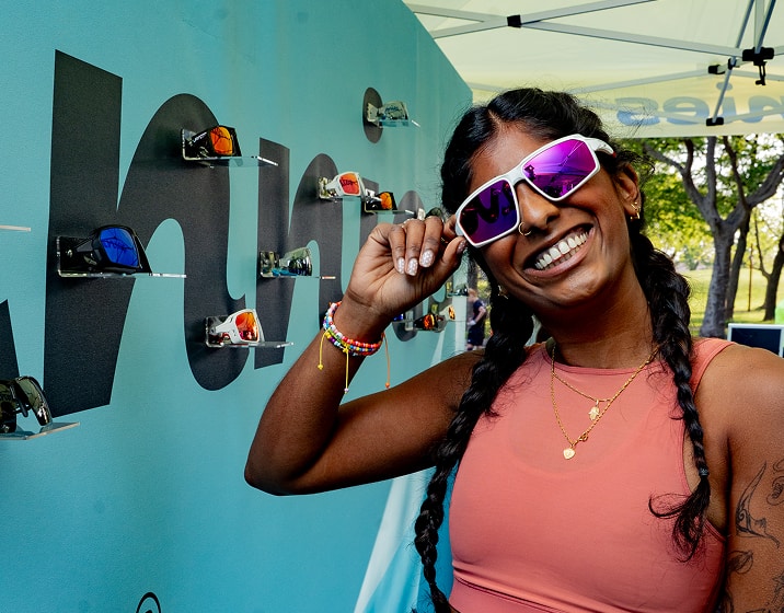 Woman posing with white framed Zunnies sunglasses at an outdoor running event, with display in the background.
