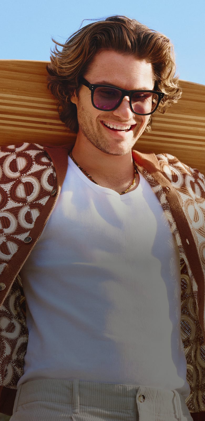 Chase Stokes in Zenni Jetsetter sunglasses, smiling outdoors in a white and brown geometric shirt against a blue sky with a wooden skateboard over his shoulders.