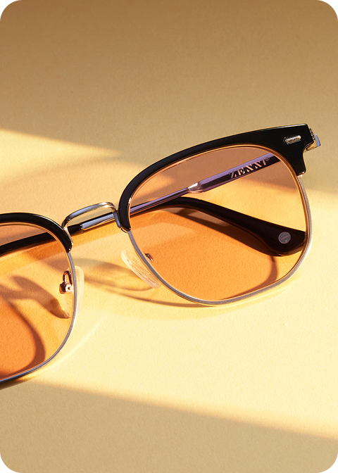 A pair of browline Game Changer Zenni x Chase Stokes sunglasses with gold accents on a light golden surface.