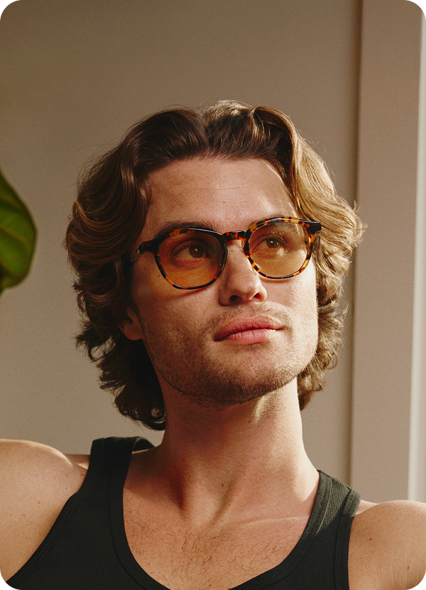 Chase Stokes sitting indoors, wearing brown Zenni Surfwatch frames with tinted lenses and a black tank top