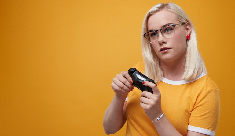 Gamer gal with shoulder-length light blonde hair wearing blue metal browline glasses #3210916 and yellow ringer tee and red heart-shaped earrings holds a game controller.