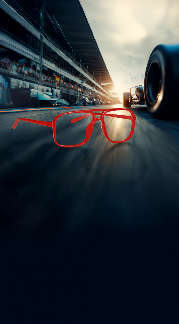 Close-up of bold red avaitor glasses on a racetrack, with a Formula 1 car speeding past and crowds watching from the pit lane in the background under a bright setting sun.