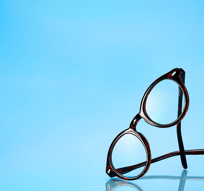 Dark tortoiseshell round eyeglasses standing upright on a reflective surface against a vibrant light blue background.
