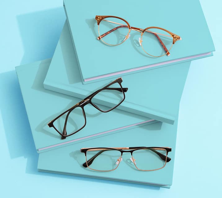 Three pairs of eyeglasses displayed on a stack of light blue books. The glasses include a round gold frame, a rectangular black frame, and a semi-rimless black frame.
