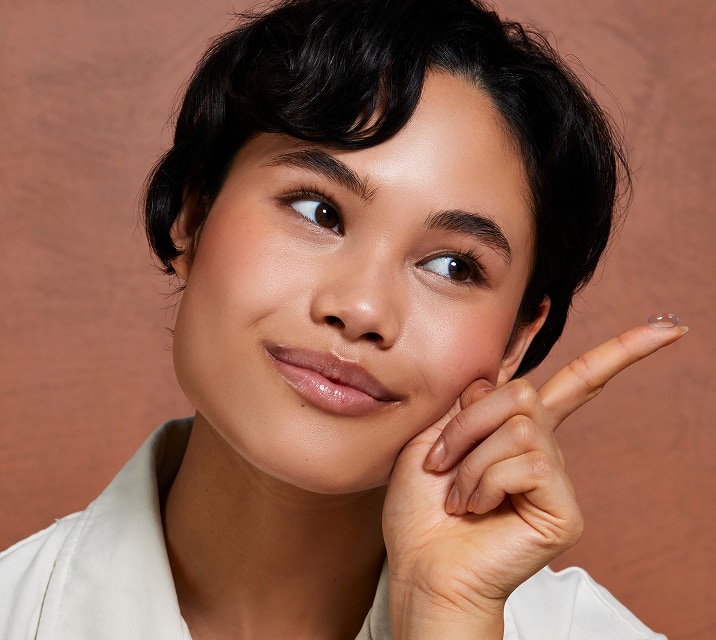 Close-up of a young woman with short dark hair, smiling and pointing to the side, holding a contact lens in her left index finger.