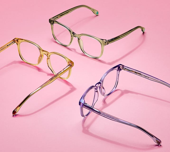Stylish transparent Zenni eyeglasses in pastel green, yellow, and purple frames displayed on a pink background, from the Zenni's Vision of Wellness collection supporting mental health.