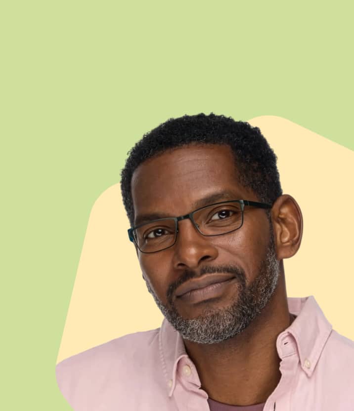 A man with a salt-and-pepper beard wearing a pink shirt and thin, dark-framed rectangular glasses with a double-bridge look, positioned against a yellow and green cloud-shaped background.