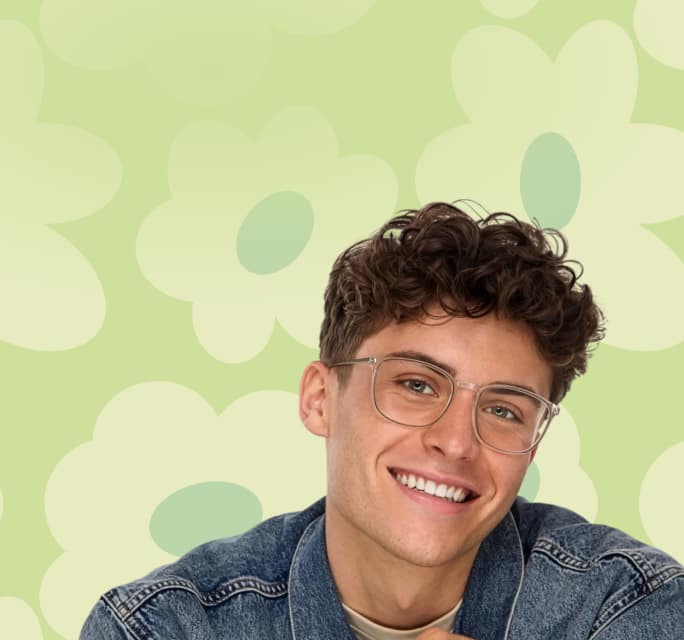 A smiling man with curly brown hair wearing translucent grey rectangular glasses and a denim jacket against a pale green background with large flower illustrations.