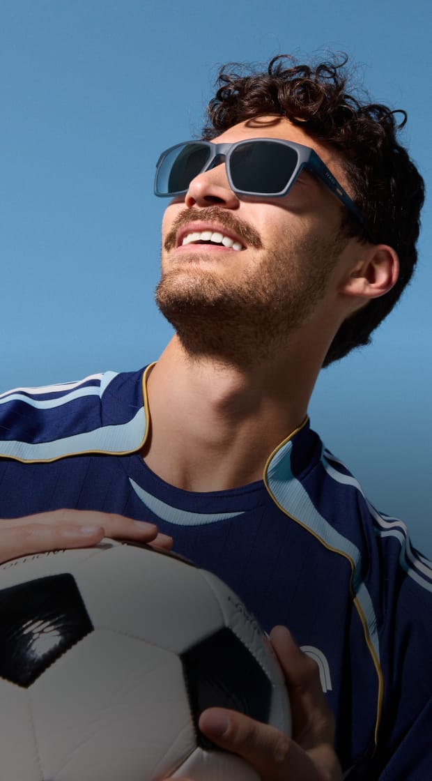Smiling man with a mustache wearing dark square sunglasses and a light blue athletic jersey, set against a clear blue sky.