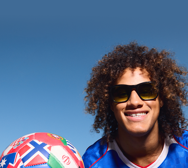 Smiling person with curly hair wearing a blue soccer jersey and black sunglasses with yellow-tinted lenses, posing with a flag-patterned soccer ball against a clear blue sky.