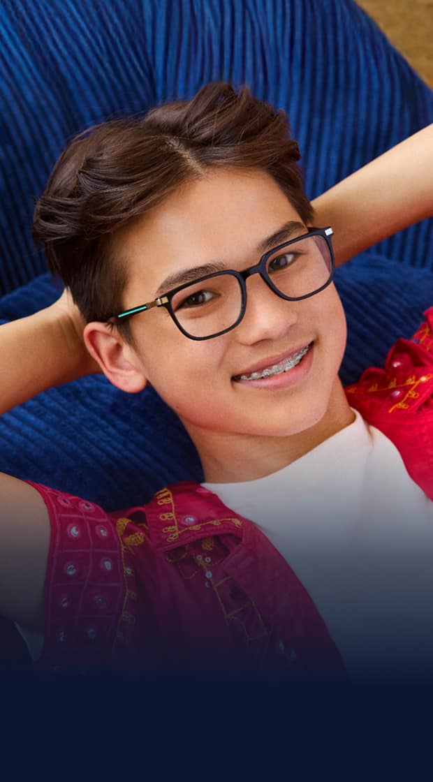 Smiling boy with short brown hair and braces wearing rectangular black-and-teal eyeglasses, a white T-shirt, and a bright pink embroidered shirt, leaning back with hands behind head against a colorful abstract background.