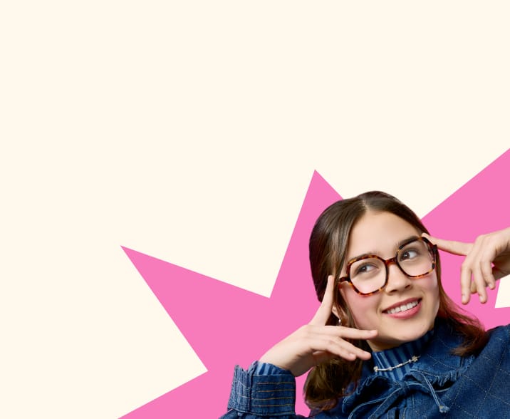 Person wearing tortoiseshell glasses and a denim outfit, smiling with fingers near their face against a pink and off-white background
