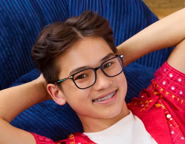 Smiling boy with short brown hair and braces wearing rectangular black-and-teal eyeglasses, a white T-shirt, and a bright pink embroidered shirt, leaning back with hands behind head against a colorful abstract background.