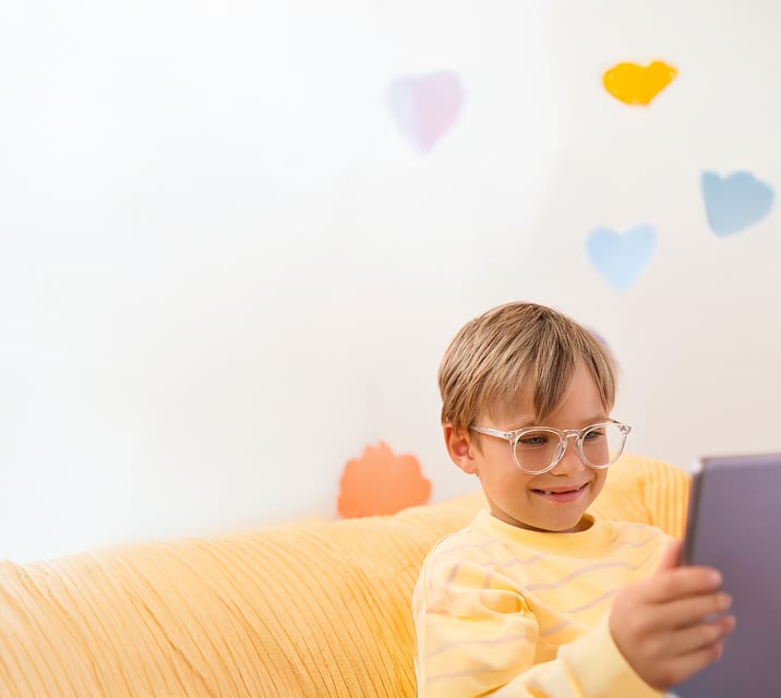 The image shows a young child with light brown hair wearing transparent glasses and a yellow sweater with pink stripes, smiling while looking at a tablet while seated on a yellow textured couch. The soft, blurred background features an orange flower shape and a blue heart shape on the wall, adding a playful touch.