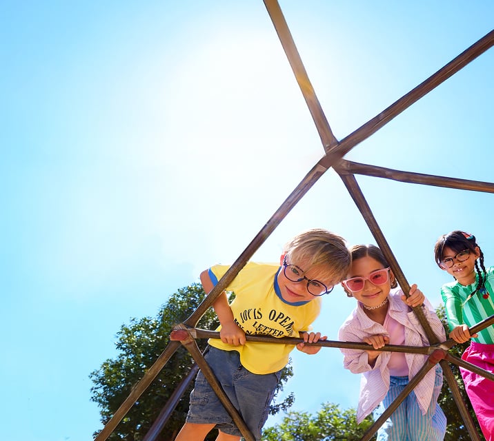 Three children wearing glasses lean on a metal play structure with a colorful backdrop.