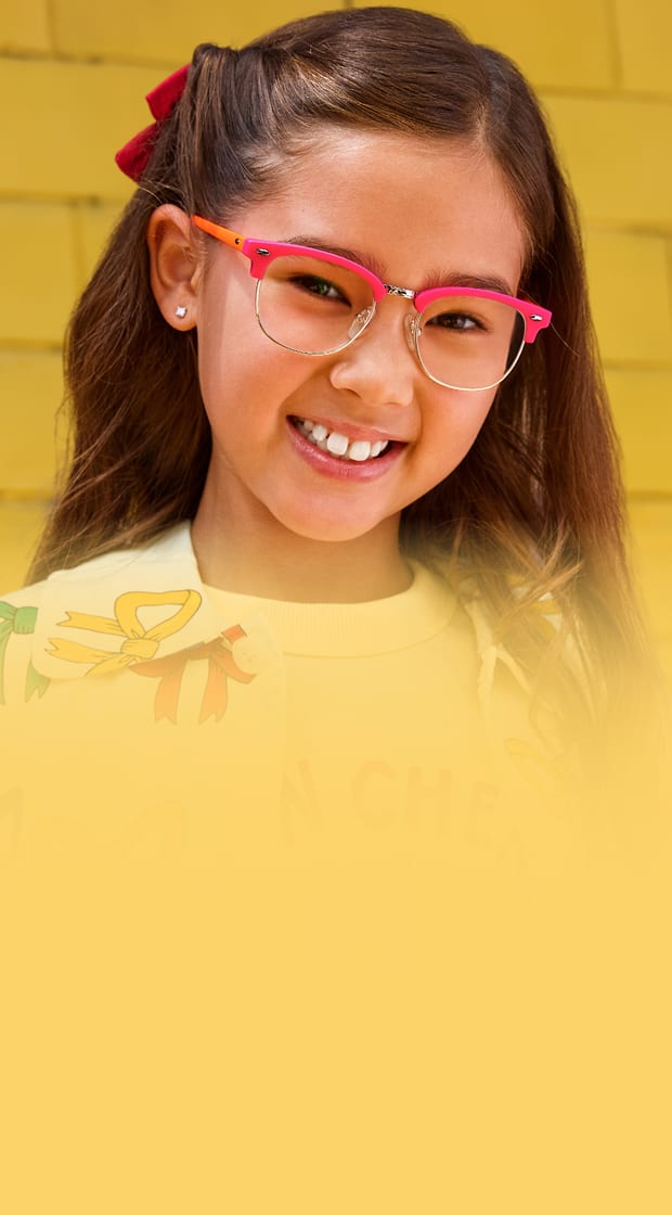 Close-up of a smiling young girl wearing bright pink and orange browline-style eyeglasses, set against a yellow brick wall background embellished with colorful hand-drawn doodle spirals and stars.
