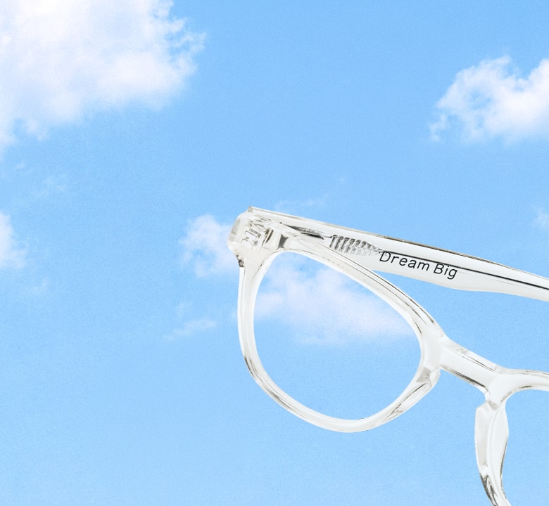 Close-up details of clear transparent round eyeglasses, showing the inscription Dream Big on the inside of the temple arm, set against a bright blue sky filled with soft white clouds.