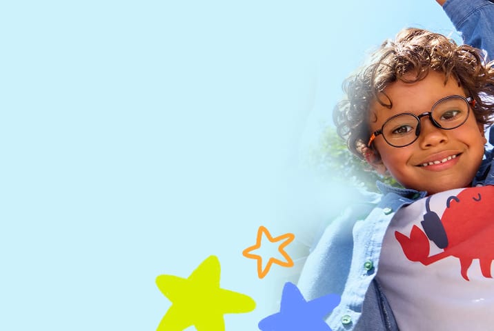 Smiling young boy with curly dark hair looking down at the camera while hanging from playground equipment, wearing round black wireframe eyeglasses and a denim shirt against a clear blue sky.