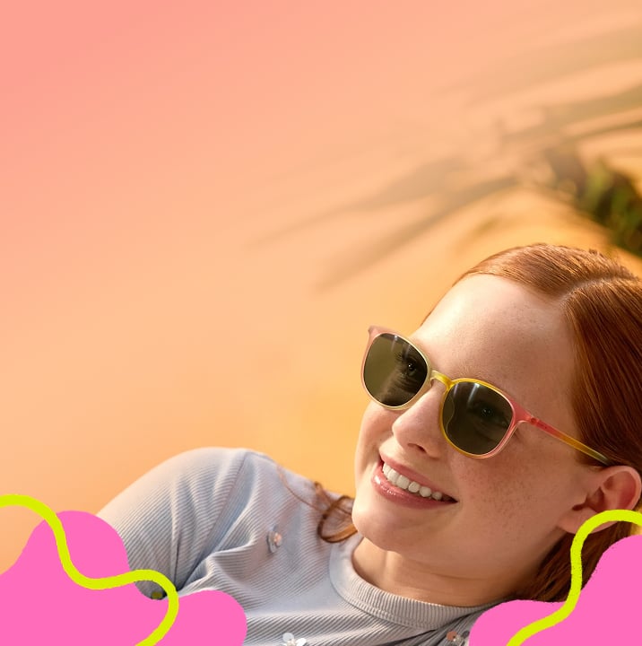 A young boy smiling outdoors wearing round-frame sunglasses with a pink-to-yellow gradient frame and dark protective lenses.