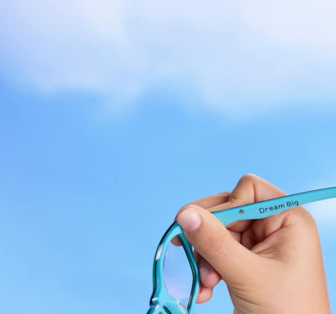 Hand holding a pair of light blue eyeglasses showing the inner temple arm engraved with the phrase Dream Big against a bright blue sky.