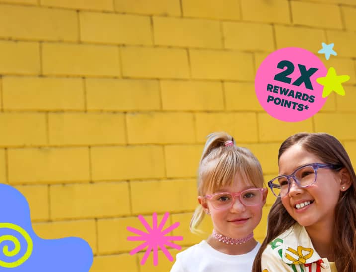 Two young girls smiling together against a bright yellow brick wall; one has blonde hair in a ponytail and wears pink rectangular glasses, while the other has dark hair and wears clear purple rectangular glasses.