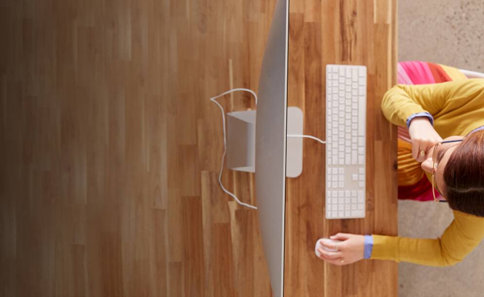 An overhead shot of a person wearing glasses sitting at a wooden desk using a computer.